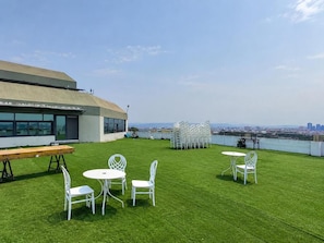 A rooftop terrace with a view of the city skyline, featuring a dining area with white chairs and tables, and a wooden bench.