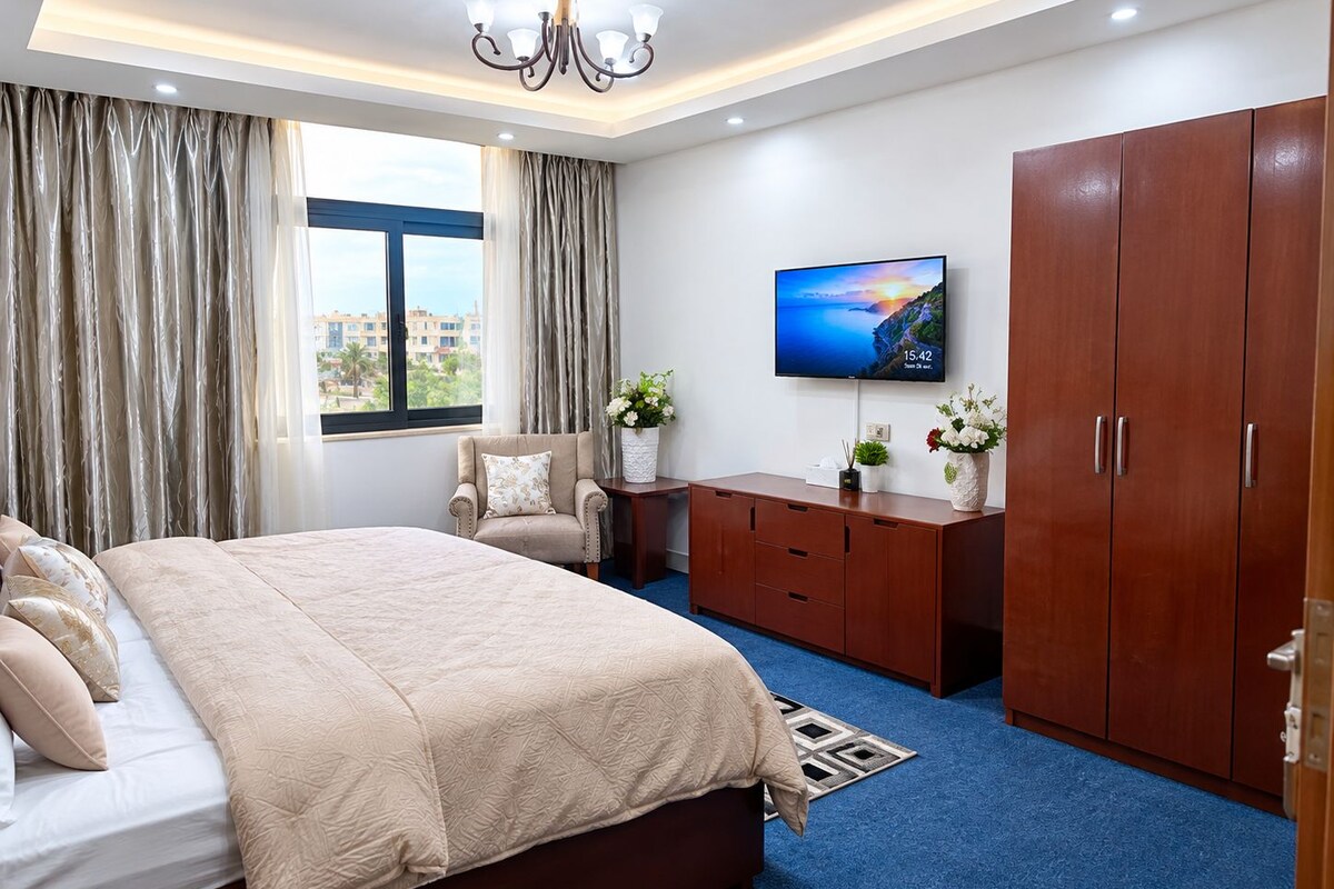 A modern hotel room with a large bed, a TV on the wall, and a view of the cityscape through the window.