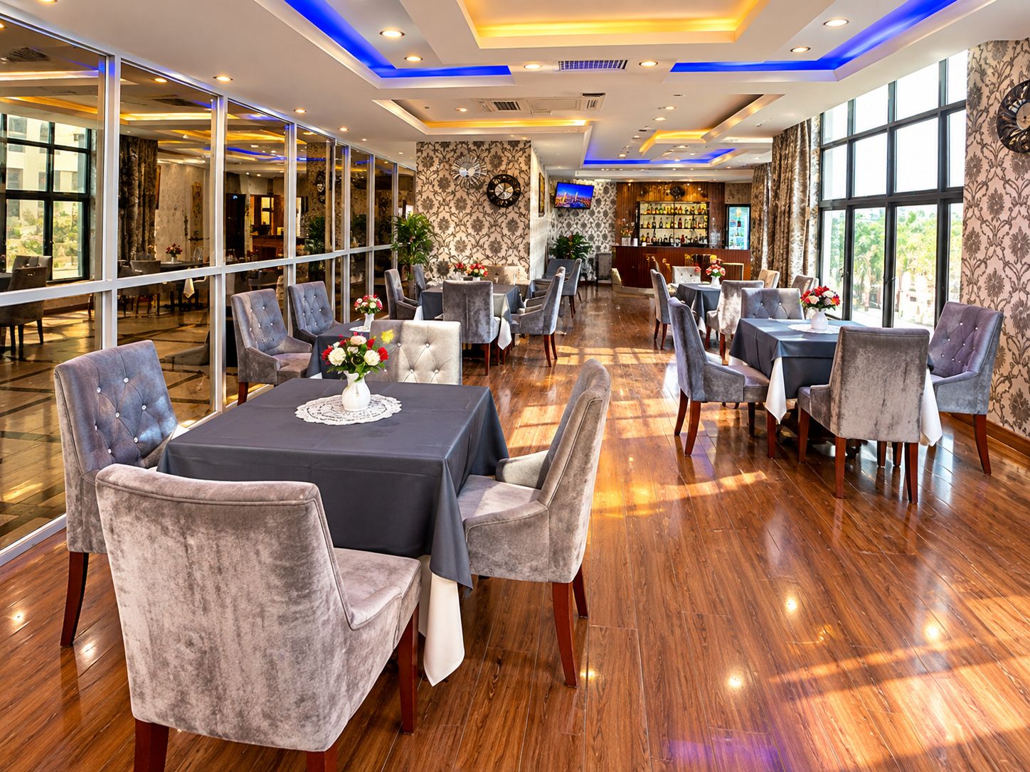 Un vaste espace dédié à la salle à manger, avec de nombreuses tables et chaises, un bar à l’arrière-plan et de grandes fenêtres laissant entrer la lumière naturelle.