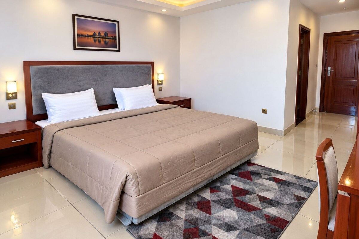 A hotel room with a large bed, a nightstand, a wall-mounted lamp, and a door.