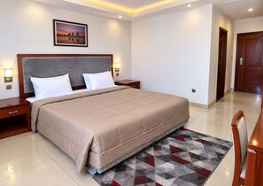 A hotel room with a large bed, a nightstand, a wall-mounted lamp, and a door. - HOTEL SAINT FRANCOIS DE PAULE  (brazzaville)