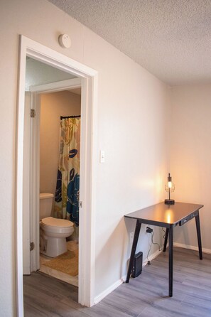 Una habitación con una puerta blanca que da a un baño, una mesita con una lámpara y suelo de madera.