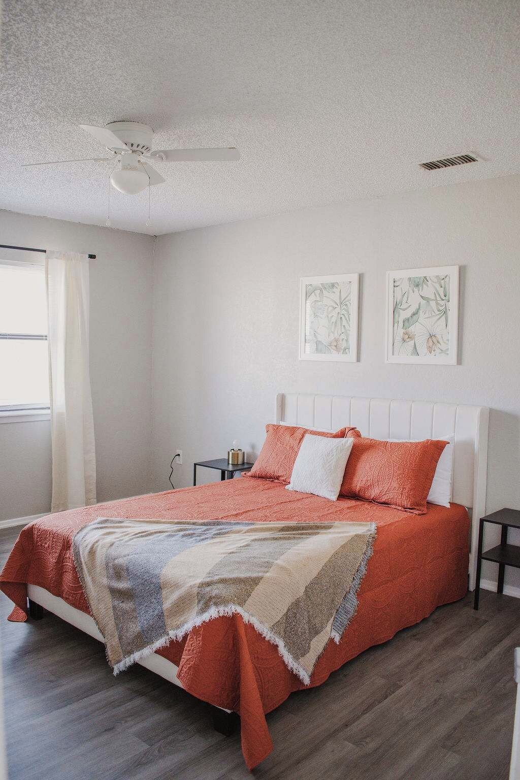 A bedroom with a bed, a ceiling fan, two wall-mounted artworks, and a window with curtains.