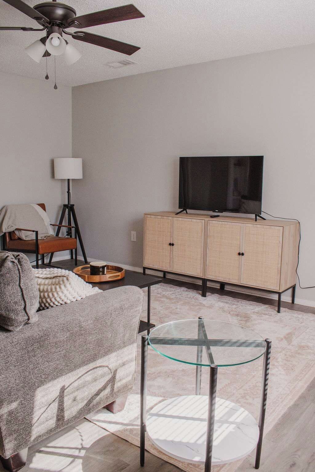 A living room with a sofa, a glass coffee table, a television on a cabinet, a floor lamp, and a ceiling fan.