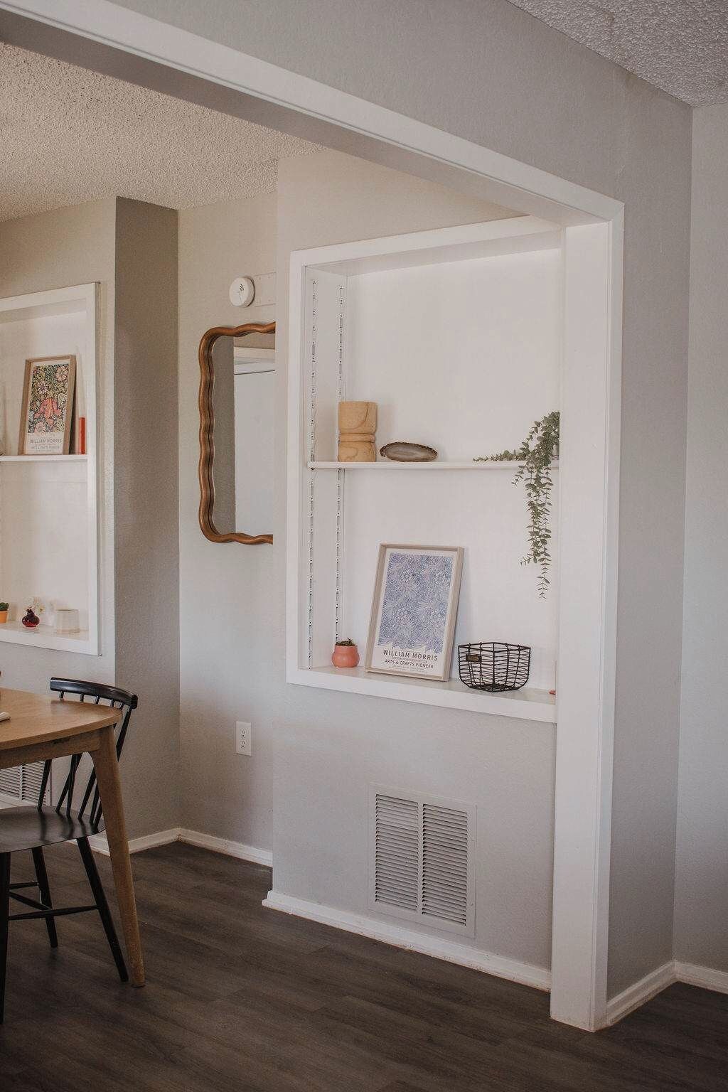 A room with a dining table, chairs, a mirror, and a wall-mounted shelf with decorative items.