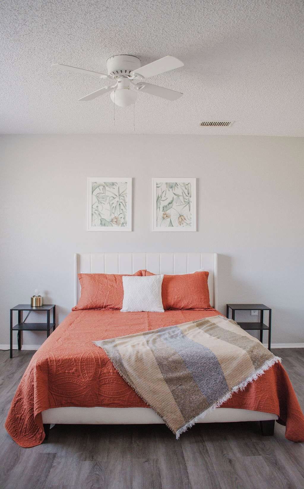 Een slaapkamer met een wit bed, twee kussens, een deken, een plafondventilator en twee ingelijste botanische prints aan de muur.