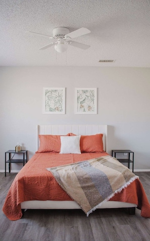 Een slaapkamer met een wit bed, twee kussens, een deken, een plafondventilator en twee ingelijste botanische prints aan de muur.