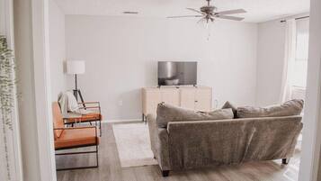 A living room with a sofa, a television on a cabinet, a ceiling fan, and a window with curtains.
