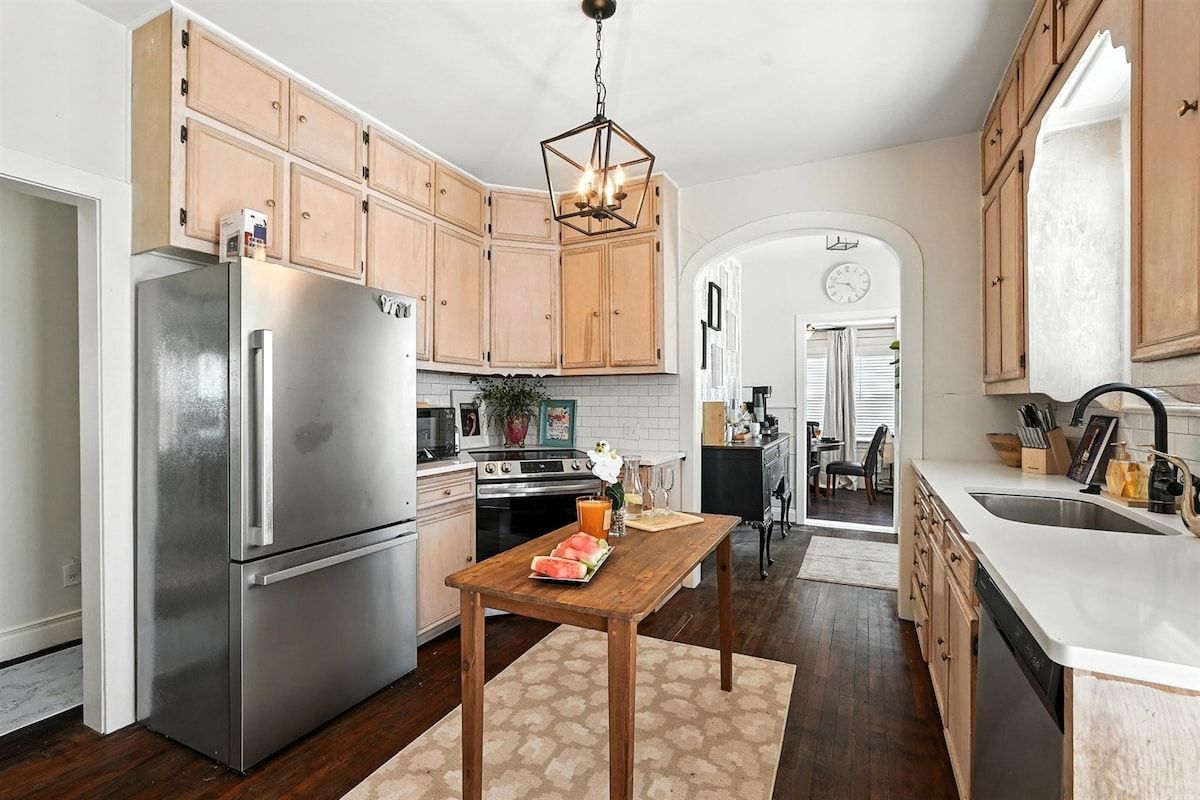 A kitchen with wooden cabinets, a stainless steel refrigerator, a wooden table with a watermelon, and a hanging light fixture.