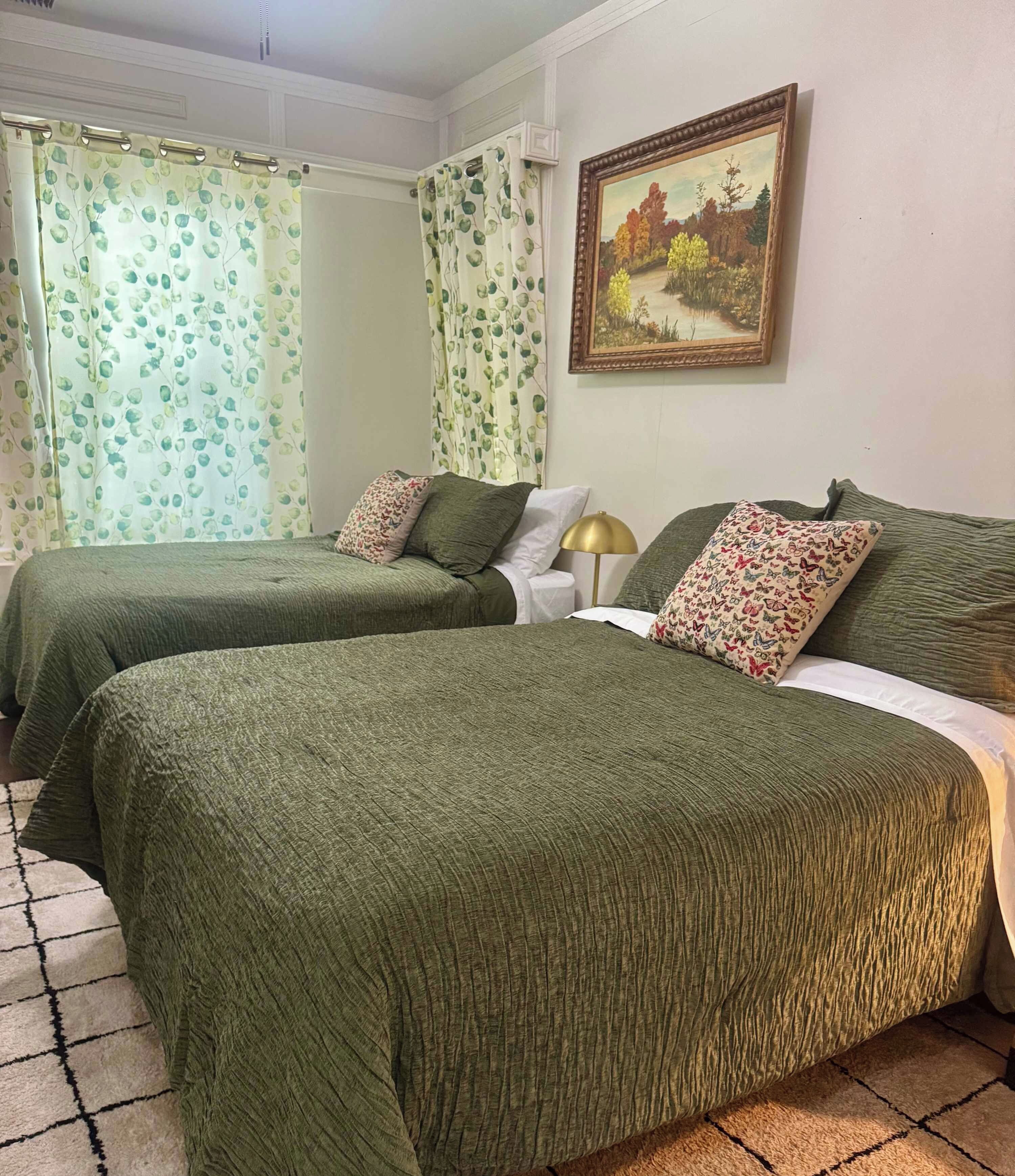 Ein Hotelzimmer mit zwei Betten, jeweils mit grüner Bettwäsche und dekorativen Kissen. An der Wand hängt ein Gemälde und es gibt ein Fenster mit Vorhängen.
