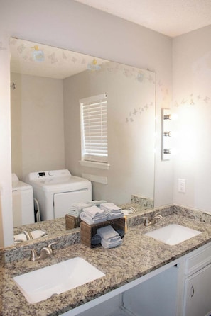 Un baño con dos lavamanos, un espejo grande, una ventana con persianas y una lavadora.