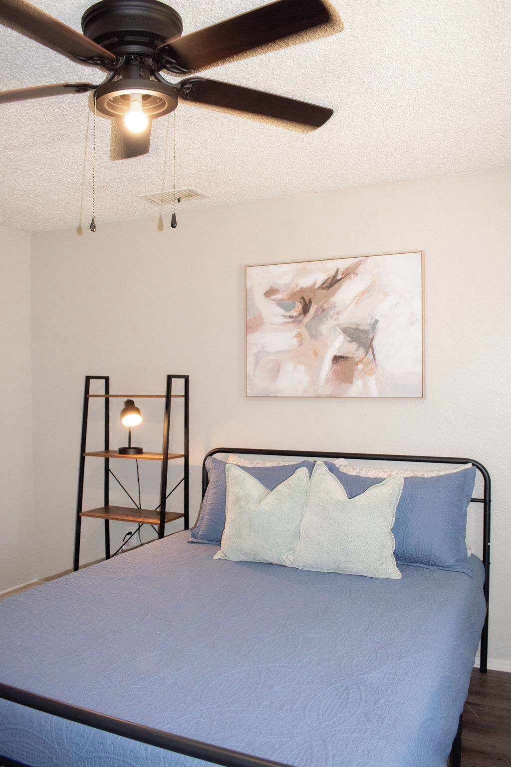 Ein Schlafzimmer mit einem Bett, einem Deckenventilator, einem an der Wand befestigten Regal mit einer Lampe und einem gerahmten abstrakten Gemälde.