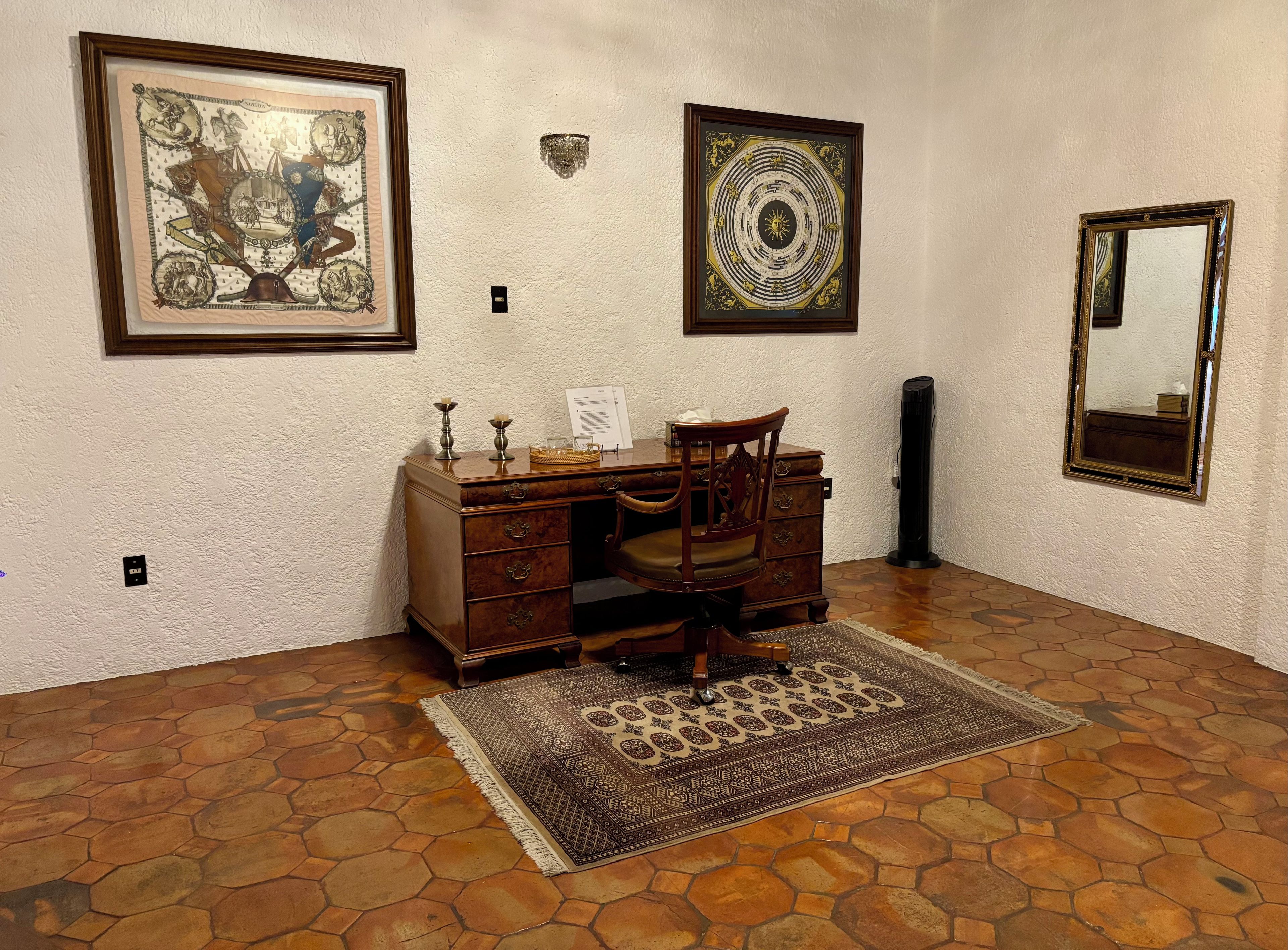 A room with a wooden desk, a chair, a patterned rug, and three framed artworks on the wall.