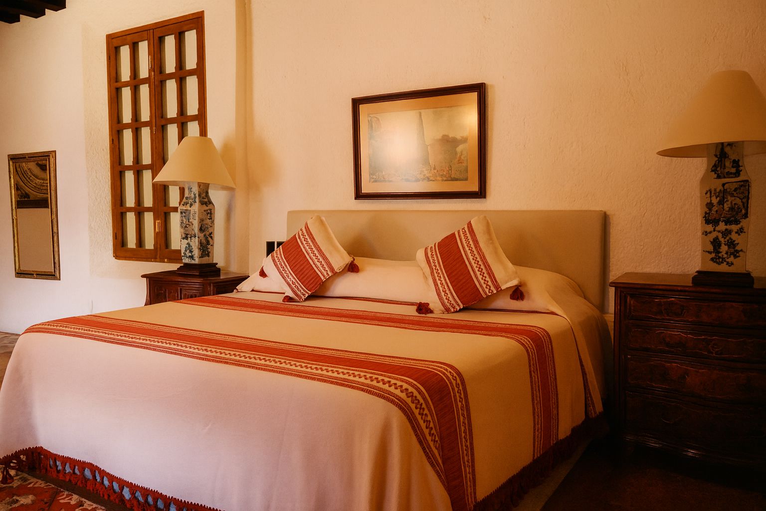 Une chambre d’hôtel avec un lit, une table de chevet, une lampe, un tableau encadré au mur et une fenêtre avec un volet en bois.