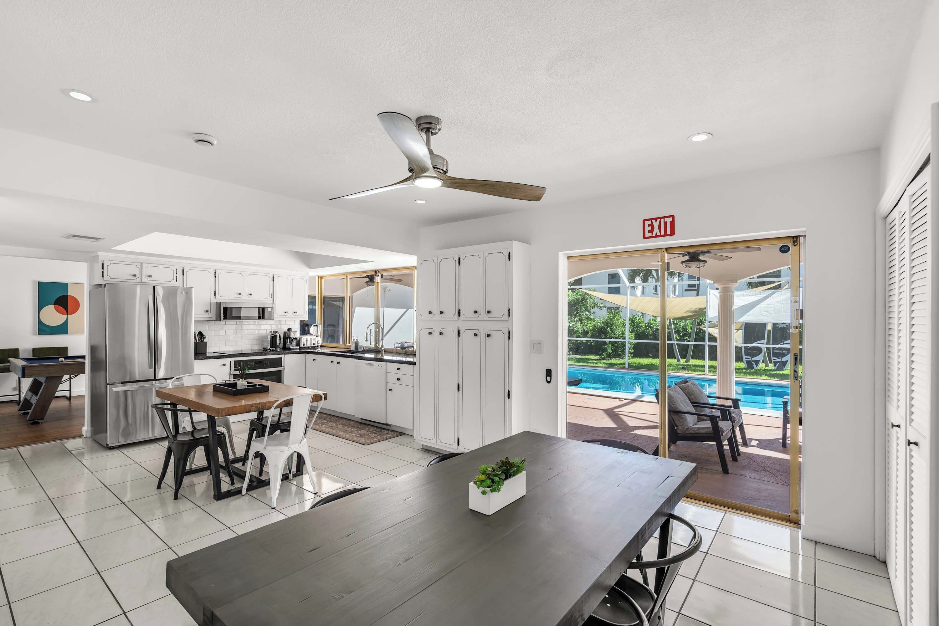 Une cuisine moderne avec des meubles blancs, un coin repas doté d’une table en bois et une vue sur une piscine, visible à travers des portes vitrées.