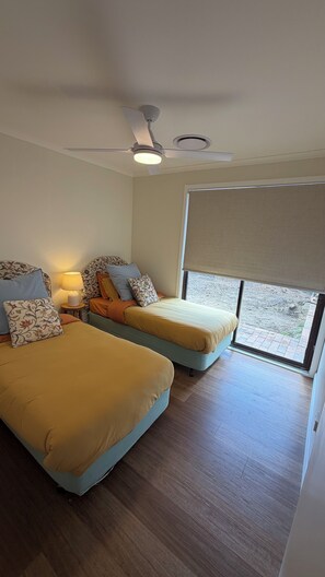 A bedroom with two beds, a ceiling fan, a window with a blind, and a nightstand with a lamp. - Elevated Central getaway in Nowra (Nowra)