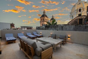Une terrasse sur le toit, avec des chaises longues, une table en bois et une vue sur des bâtiments historiques se détachant sur le ciel au coucher du soleil.