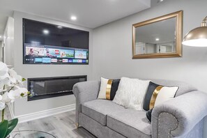 Ein Wohnzimmer mit einem grauen Sofa, einem an der Wand befestigten Flachbildfernseher, einem großen Spiegel und einer dekorativen Lampe.