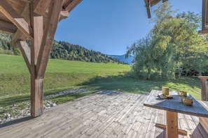 A wooden deck with a table and chairs, overlooking a green field and forested hills. - Chalet Nejma - Welkeys (Megève)
