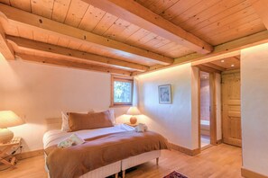 A cozy bedroom with a wooden ceiling, a bed with a brown bedspread, a bedside table with a lamp, and a window with a view of trees. - Chalet Nejma - Welkeys (Megève)