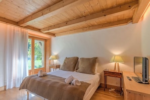 A bedroom with a wooden ceiling, a bed with a brown blanket, two bedside lamps, a nightstand with a lamp, and a TV on a wooden table. - Chalet Nejma - Welkeys (Megève)