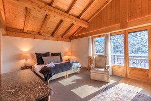 Quarto aconchegante com cama, abajures, uma cadeira e vista para uma paisagem nevada através de grandes janelas.