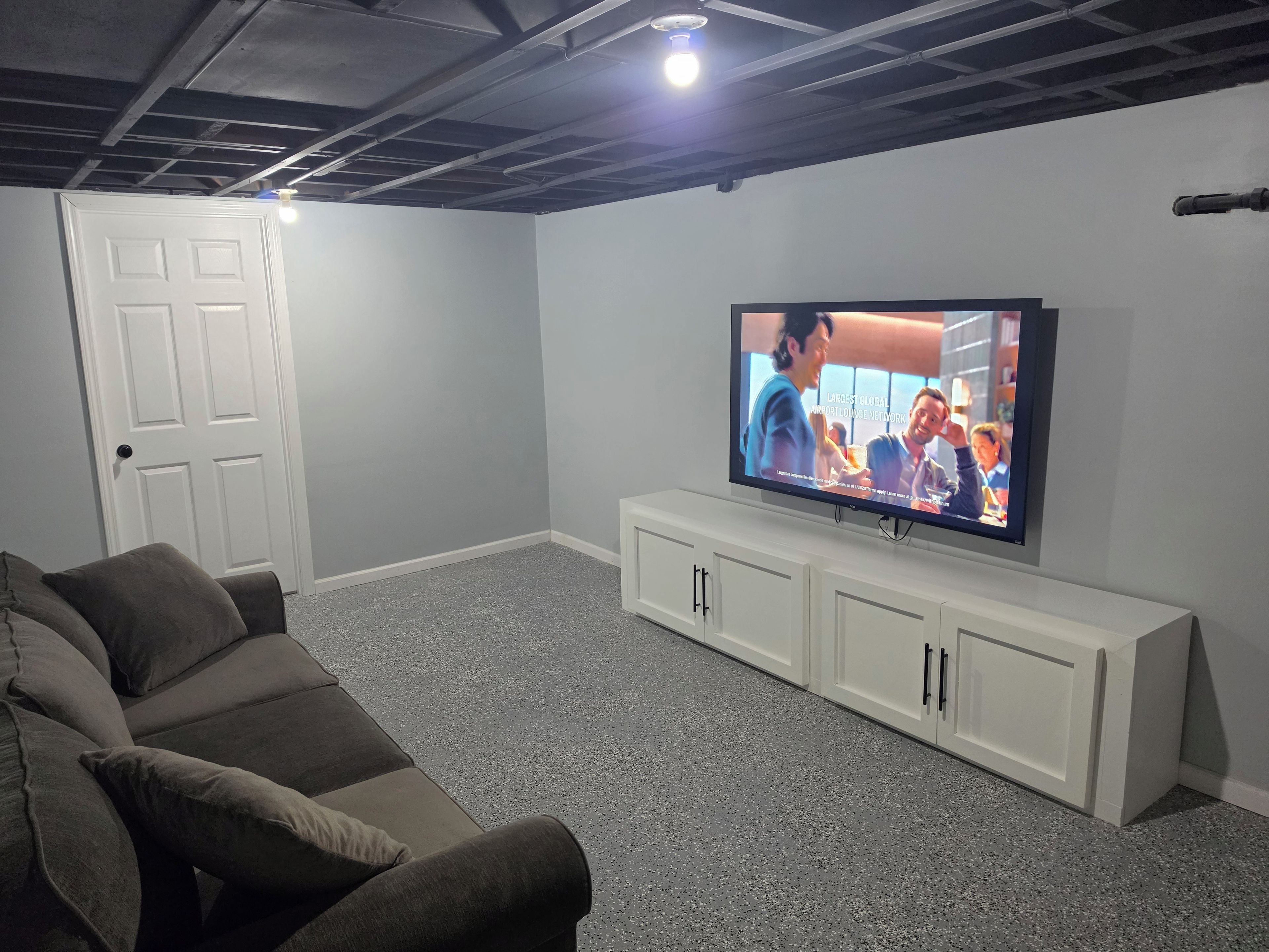 A room with a television on a white stand, a grey speckled carpet, a grey sofa, and a white door.