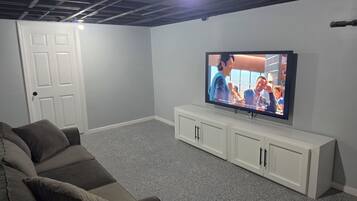A room with a television on a white stand, a grey speckled carpet, a grey sofa, and a white door.