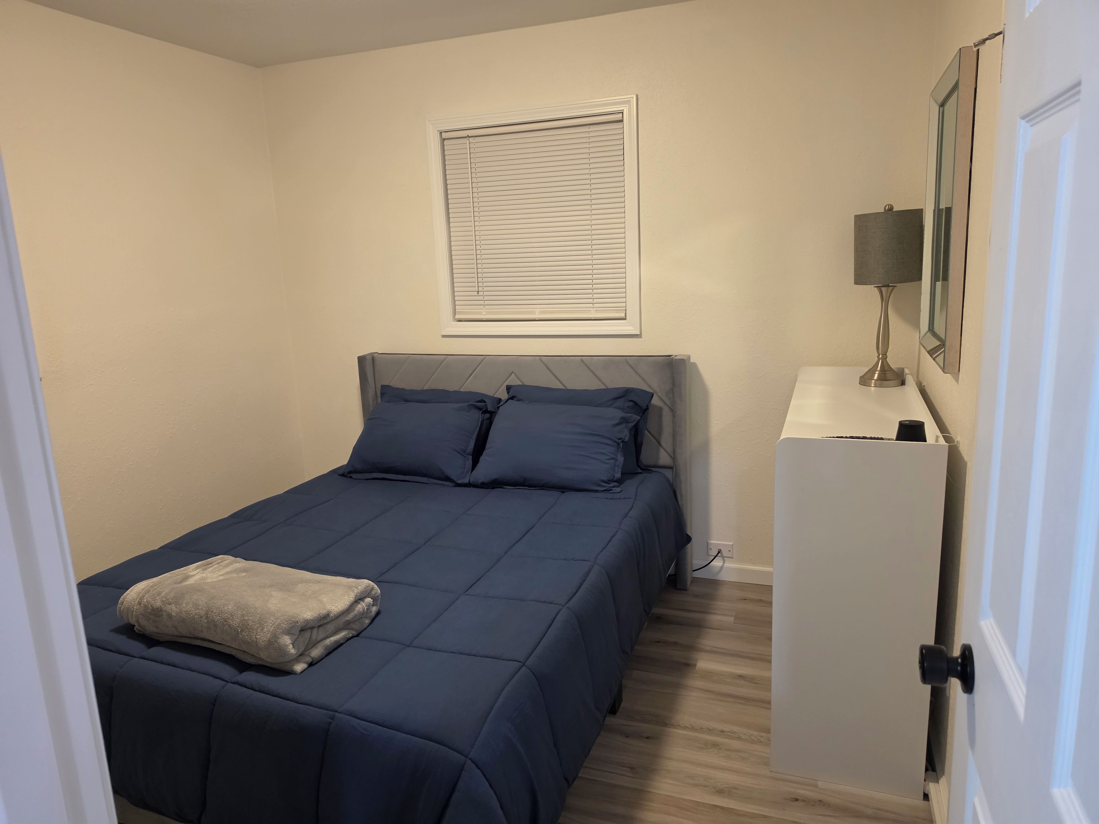 Un dormitorio con cama, mesita de noche con lámpara, un cuadro en la pared y una puerta.