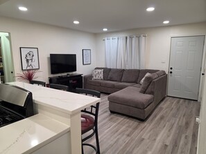 Een moderne woonkamer met een hoekbank, een flatscreen televisie, een eettafel met stoelen, en een keuken met een fornuis.