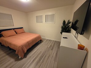 Een slaapkamer met een bed, een televisie, een plant en twee ramen met zonwering.