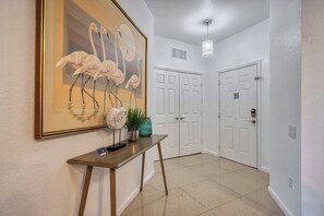 Un couloir orné d’un tableau encadré représentant des flamants roses, une console sur laquelle sont disposés des objets décoratifs, une porte blanche avec un panneau « interdiction de fumer » et un luminaire suspendu.