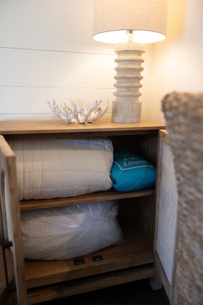 A wooden nightstand with a lamp, a decorative coral piece, and storage compartments with folded linens. - The Peachy Palm | St. Simons Island Condo Near Pier Village (Saint Simons Island)