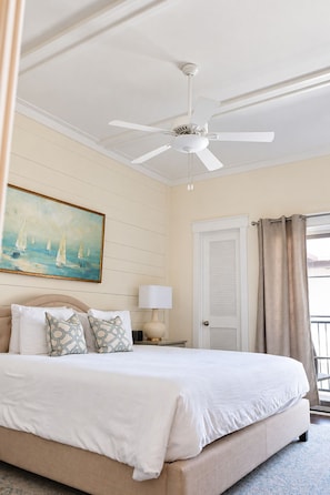 A hotel room with a bed, a ceiling fan, a painting, a nightstand, a lamp, a door, and a view of the outside. - The Peachy Palm | St. Simons Island Condo Near Pier Village (Saint Simons Island)