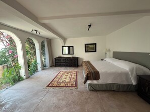 A bedroom with a large bed, a dresser, a mirror, and a window with a view of greenery.