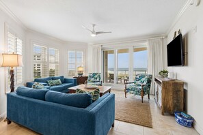 A spacious living room with a blue sofa set, a ceiling fan, and a view of the ocean through large windows. - Beautiful oceanfront condo on the quiet southend of Amelia Island (Fernandina Beach)