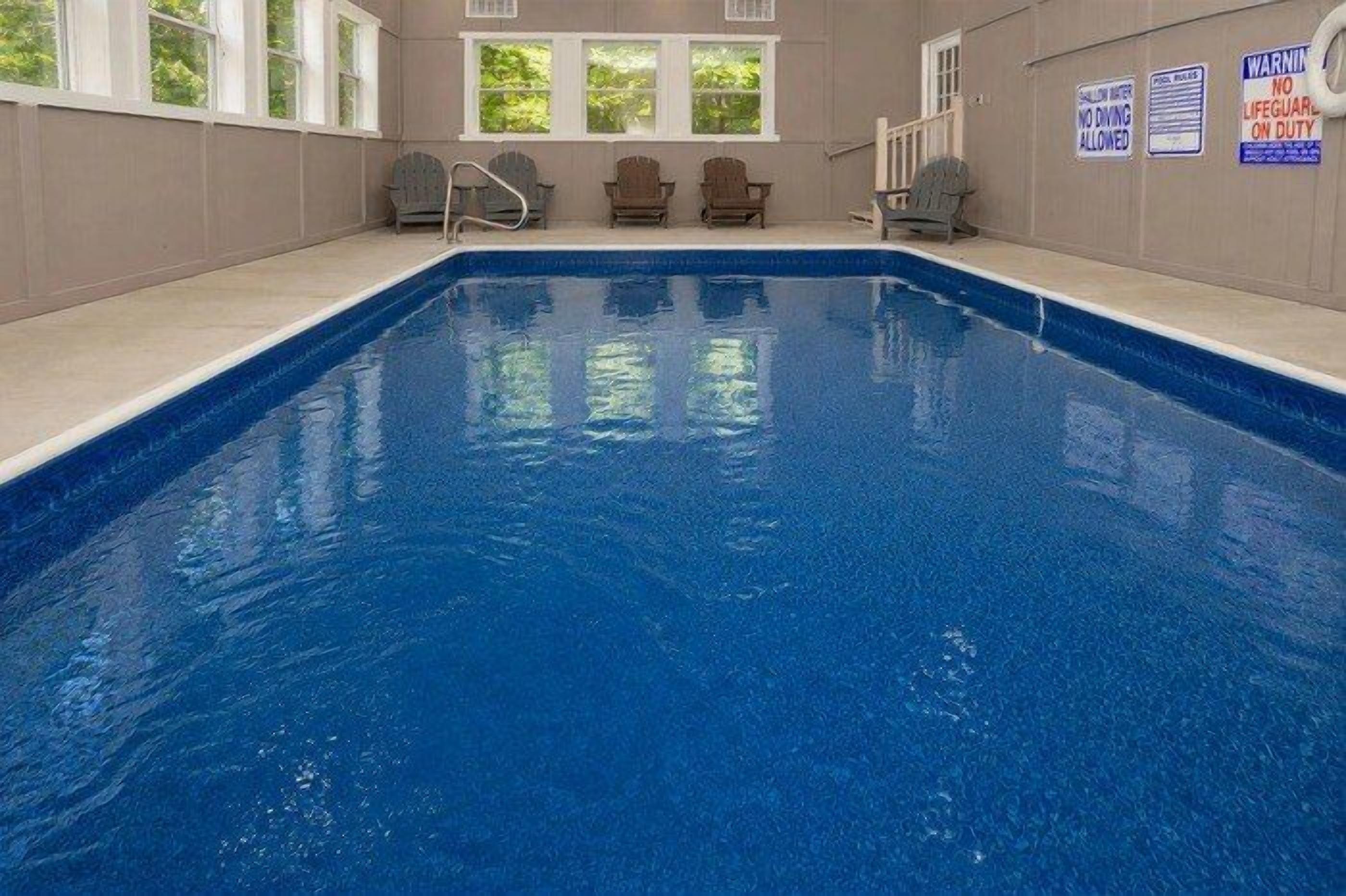 An indoor swimming pool with a clear blue water surface, surrounded by windows and a few chairs.