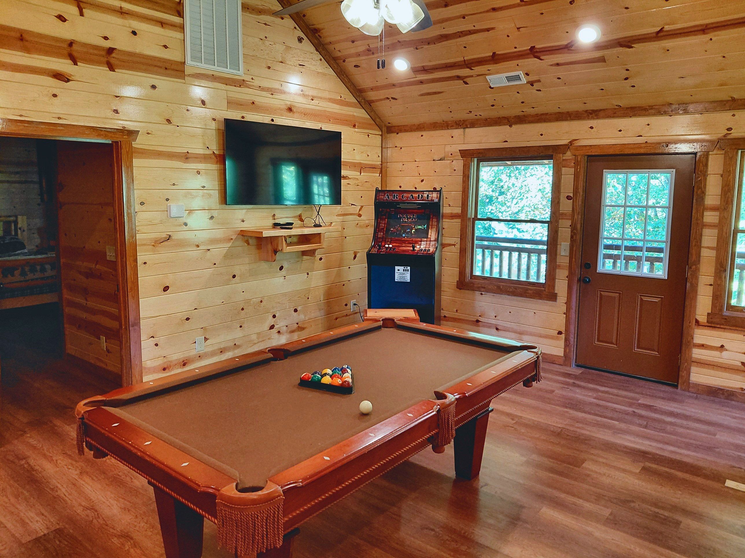 A wooden interior with a pool table, a television mounted on the wall, and a door leading outside.