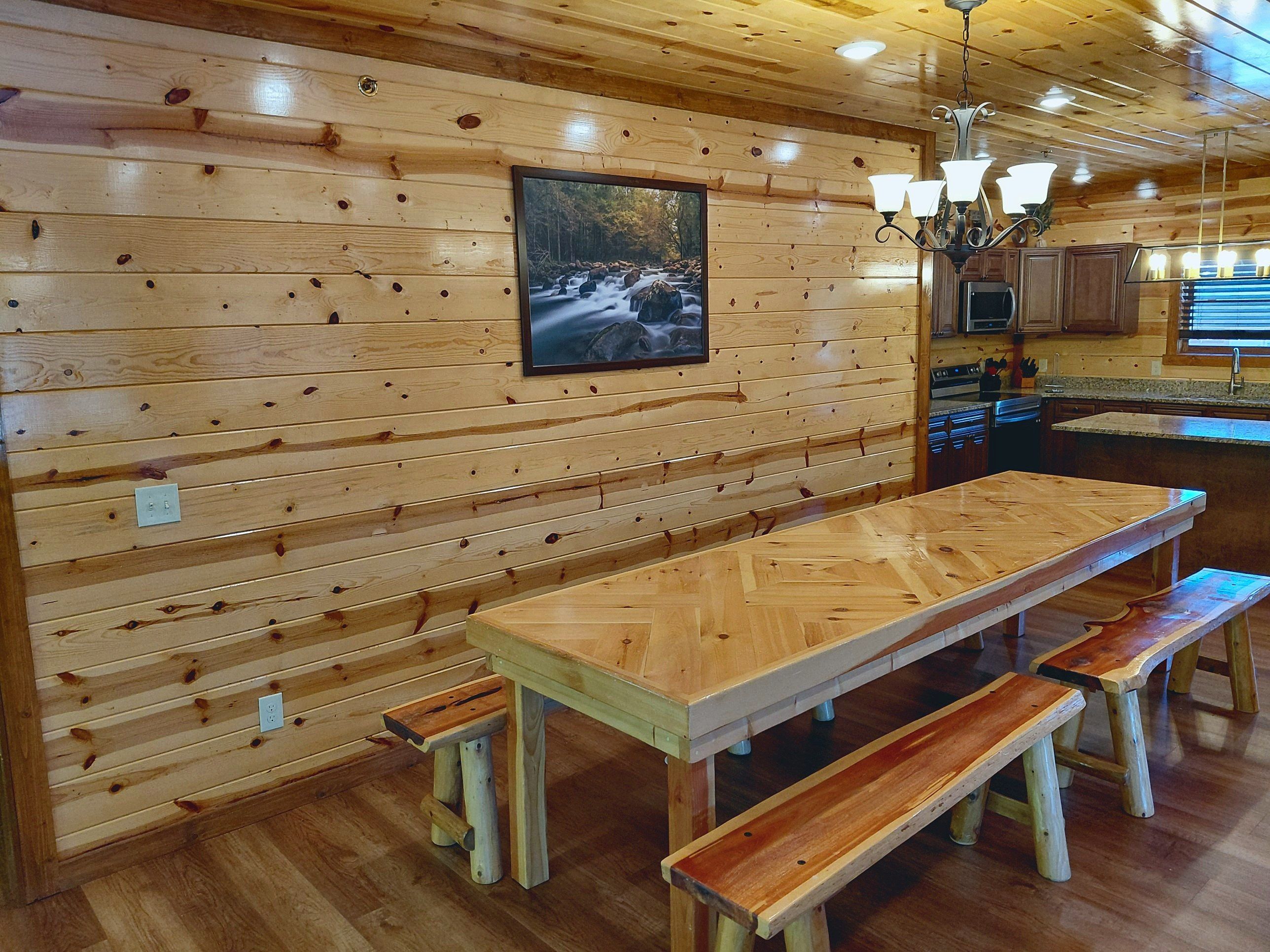A wooden interior with a long dining table and benches, a kitchen area, and a framed picture on the wall.
