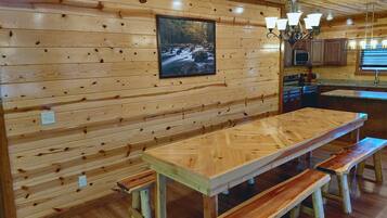 A wooden interior with a long dining table and benches, a kitchen area, and a framed picture on the wall.