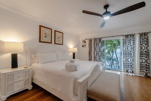 A bedroom with a bed, two bedside lamps, a ceiling fan, a window with patterned curtains, and artwork on the wall. - Evie Mar | Keys Waterfront Escape + Pool & Dock & Kayak & Paddle board & Fishing (Marathon)
