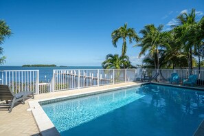 A swimming pool with a view of the ocean, surrounded by palm trees and a white fence. - Evie Mar | Keys Waterfront Escape + Pool & Dock & Kayak & Paddle board & Fishing (Marathon)