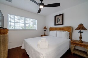 Habitación de hotel con una cama, ventilador de techo, ventana con persianas, dos lámparas de noche y un cuadro en la pared.