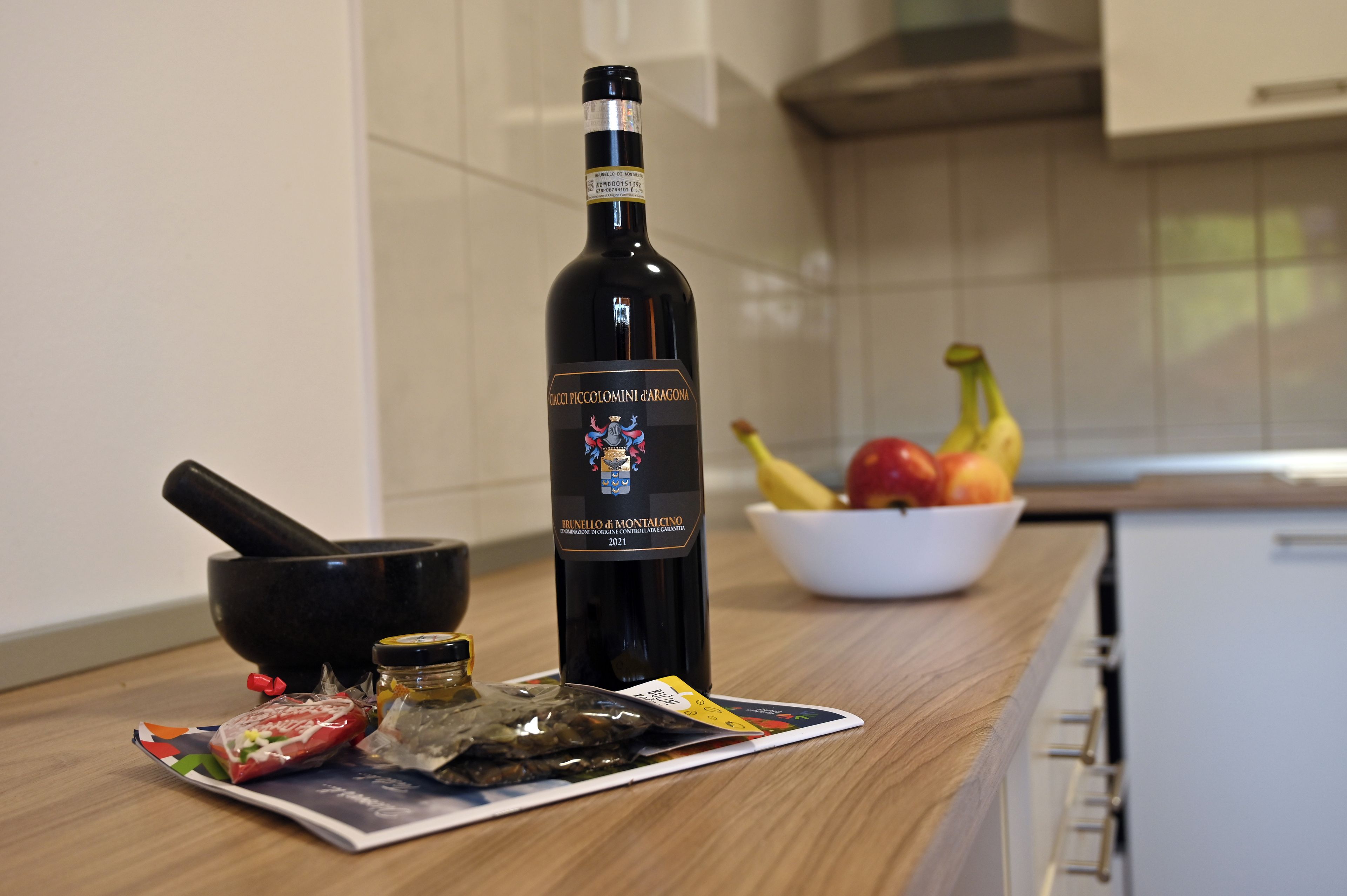A bottle of Brunello di Montalcino wine on a kitchen counter with a bowl of fruit and a magazine.