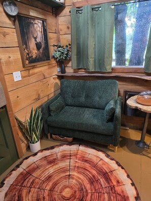 A cozy room with a green sofa, a wooden wall clock, a framed deer picture, a potted plant, and a round wooden table.