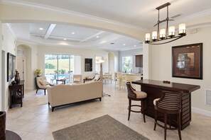 A spacious living area with a sofa, dining table, and chairs, a kitchen area, and large windows offering a view of the outdoors. - Serene University Park Golf & Country Club Pool Home - Gulf Standard (Bradenton)