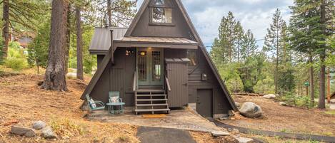 A cabin with a steeply pitched roof, a front porch with a set of stairs, and a small outdoor seating area.