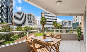 En balkon med et træbord og stole, der udover et bybillede med moderne bygninger og grønt.