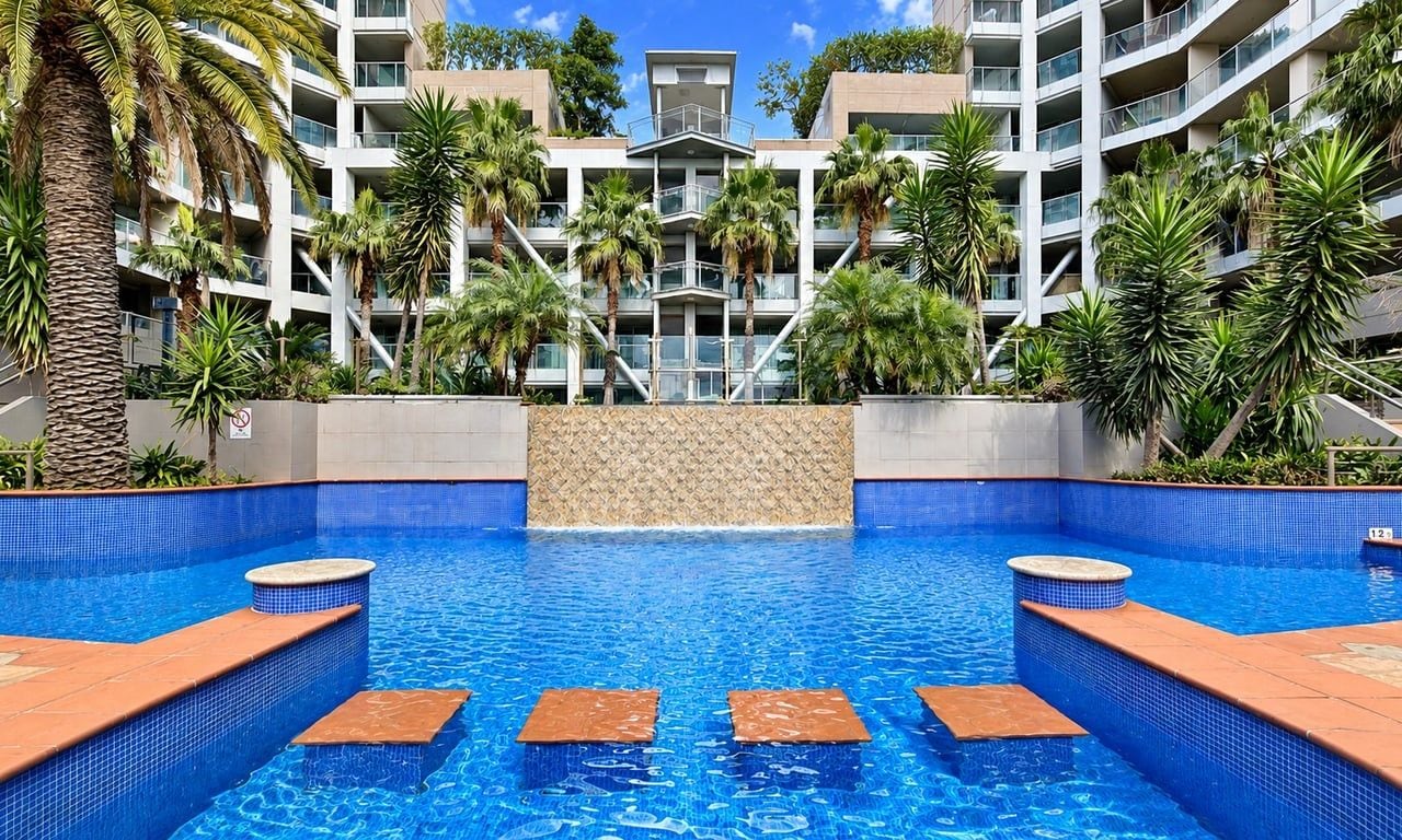 A swimming pool with steps leading into it, surrounded by palm trees and a building with multiple balconies.