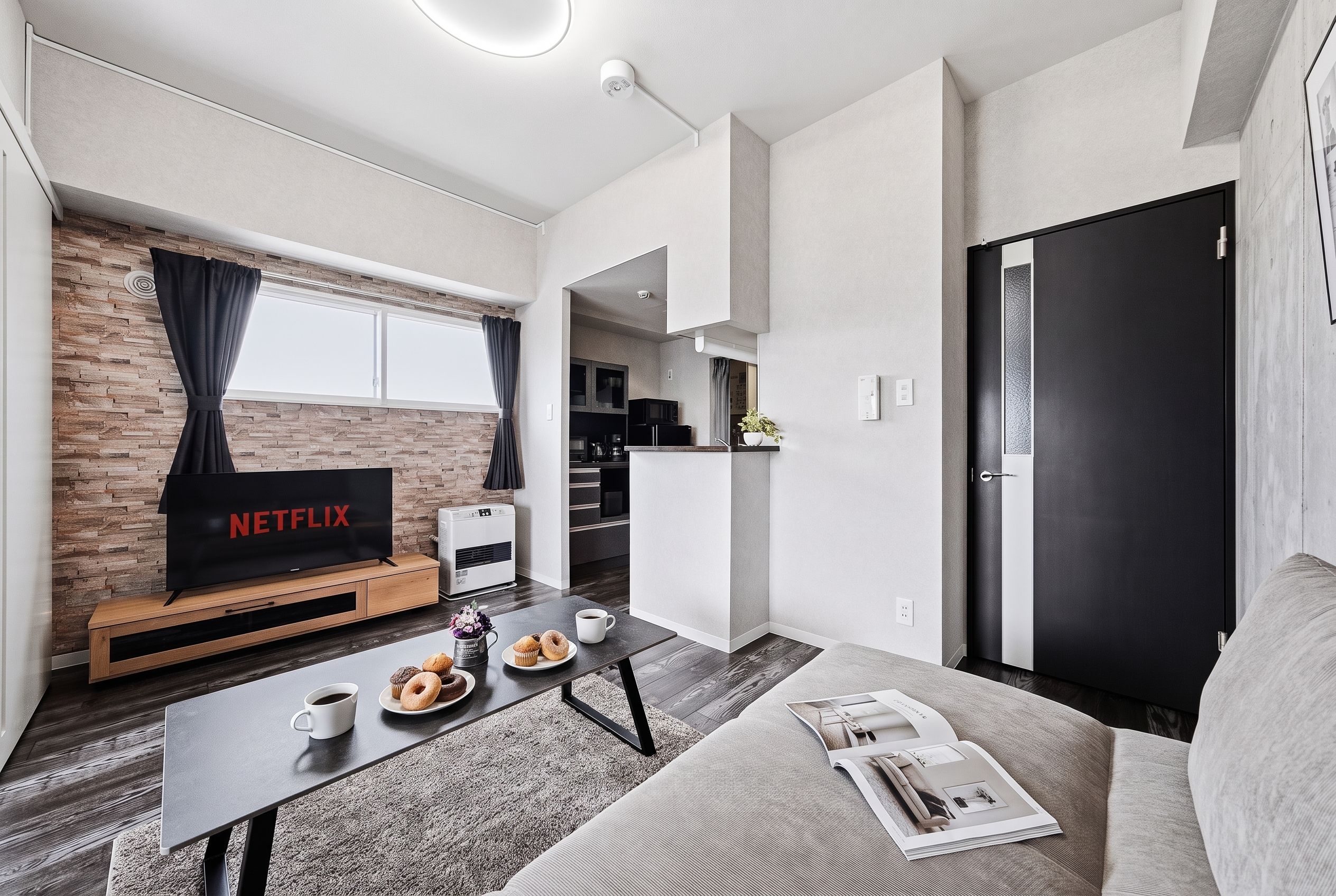 Un espacio habitable moderno con un televisor de pantalla plana, una mesa de centro con pasteles, un sofá y una cocina visible a través de una puerta abierta.
